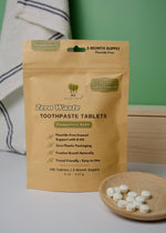 Load image into Gallery viewer, Package of zero waste toothpaste tablets with a wooden dish of tablets on a light surface.
