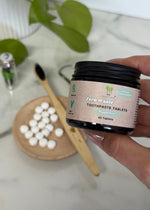Load image into Gallery viewer, Hand holding a container of zero waste toothpaste tablets with a wooden toothbrush and tablets on a white surface.
