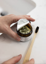 Load image into Gallery viewer, Person holding a tea bag over a jar of green capsules on a white countertop.
