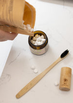 Load image into Gallery viewer, Person pouring items into a container with a bamboo toothbrush and cork lid on a white surface.

